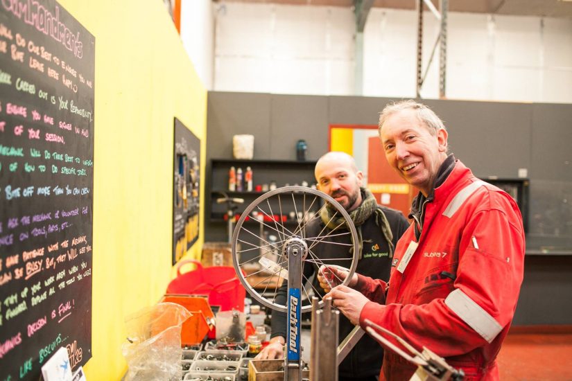 Two men are stood fixing a bicycle wheel. They face the camera and are smiling. The person on the left wears a Bike for Good staff jumper and the person on the right wears a red jumpsuit. They are stood in a mechanics workshop next to a black board.