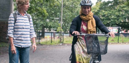 Two women outside - one is wearing a hijab with a helmet and she is on a bike that has a basket. The other is wearing a striped top and is looking at the rider