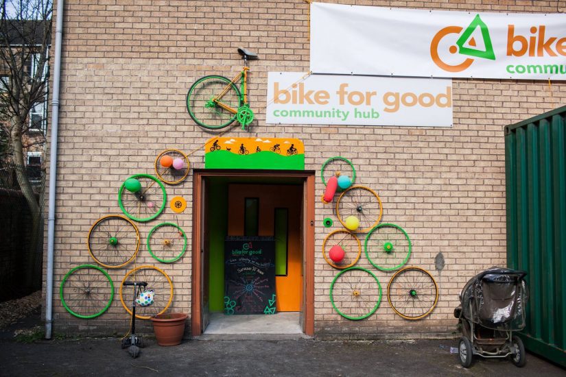 Brightly coloured bicycle wheels are attached to the side of a building around an open door frame. Above the door a large sign says bike for good community hub. On the left, a bike pump and plant pot are sat on the floor. A child's push chair is on the right.