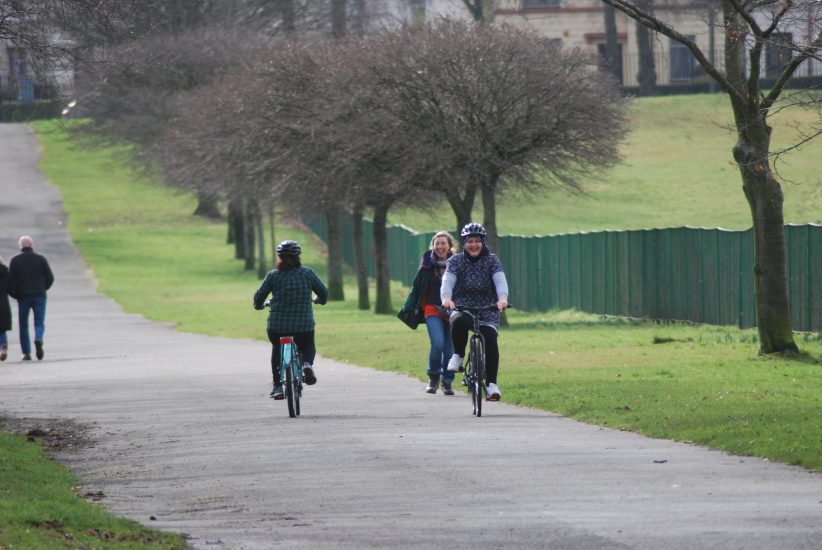 Two people are riding bicycles in a city park. One is cycling towards the camera and one cycles in the opposite direction. They are both wearing bicycle helmets.