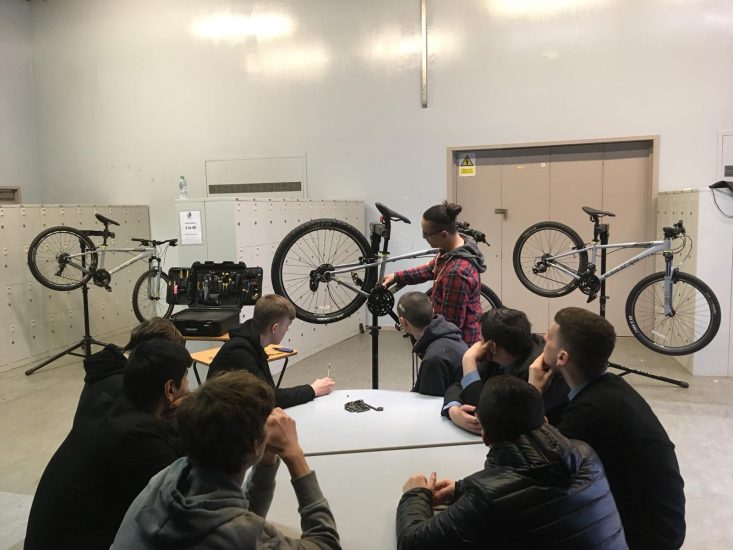 A group of young people are sat indoors around a table as one person is stood next to a bicycle. Those sat around the table are watching the bicycle.