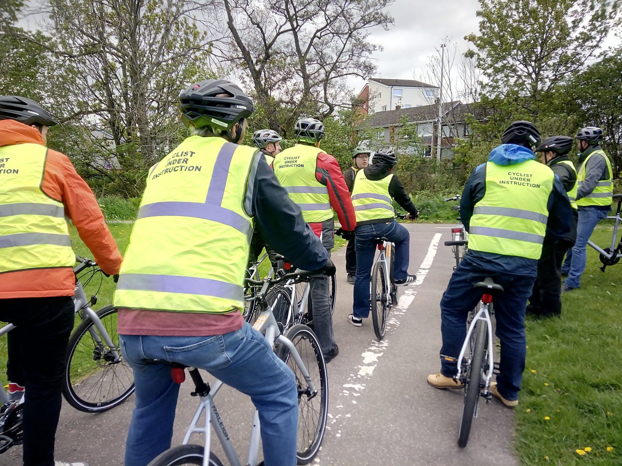 A group of people are mounted on bicycles facing away from the camera outdoors on a bike path. They each are wearing bicycle helmets and hi vis jackets with cyclist under instruction printed on the back.