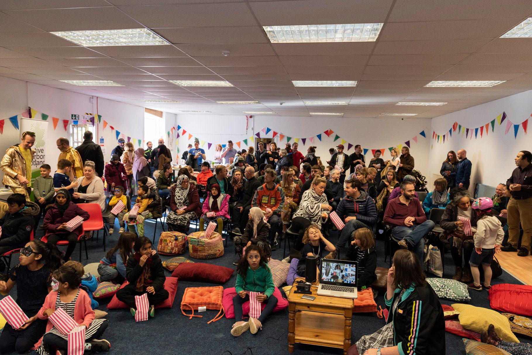 A large room is full of people of different ages, people are sat on the floor, as well as being stood or sat on chairs. In the foreground young children are eating popcorn out of paper bags. Around the room decorative bunting is hung on the walls. 