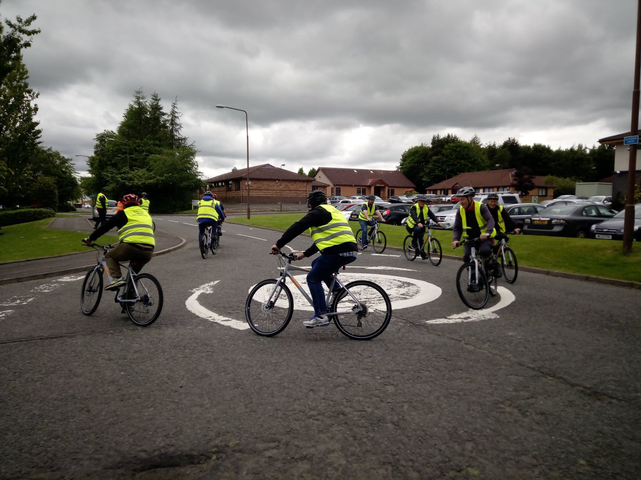 A group of people riding bicycles are cycling across a roundabout. The each are wearing hi vis jackets and are in a residential area.